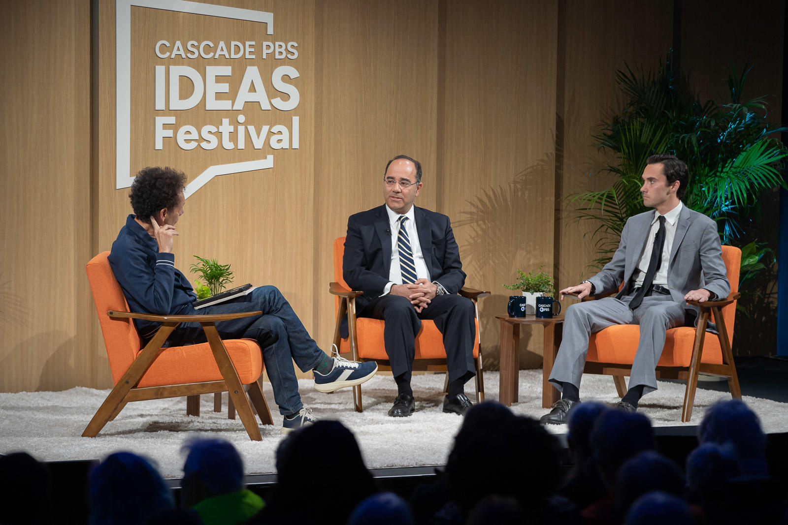 Three men sit on orange chairs on the Cascade PBS Ideas Festival stage