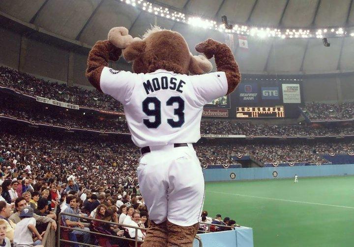 Mariner Moose is seen from behind as he flexes in front of a crowd at a baseball stadium