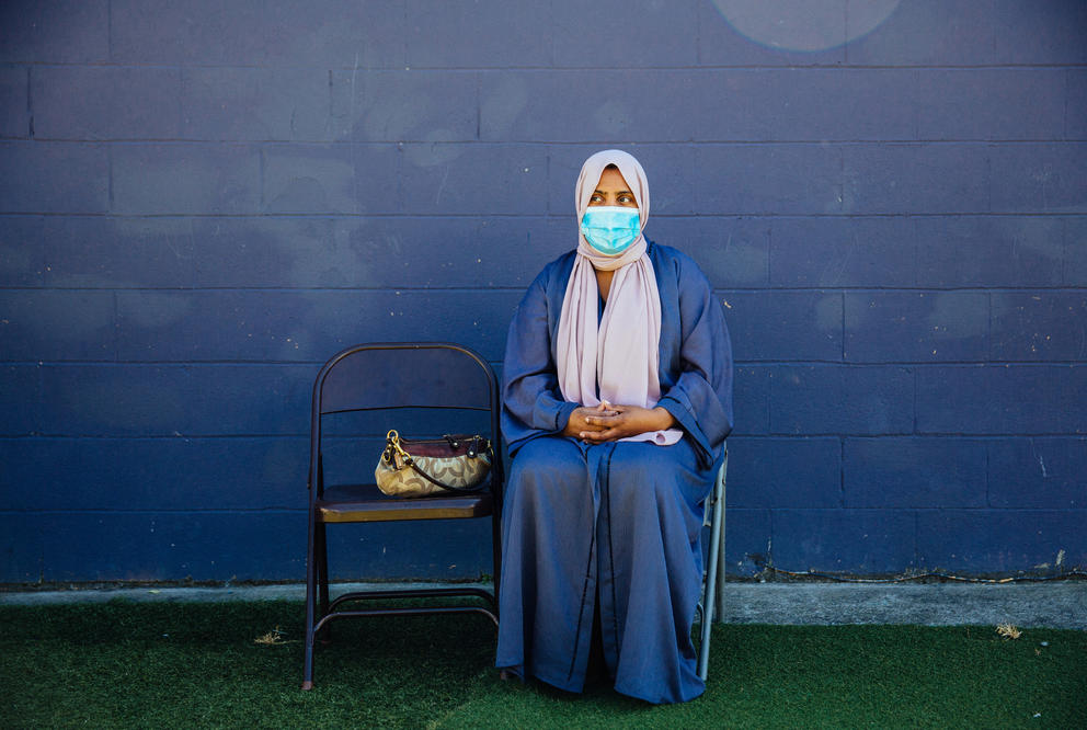 Hyatt Ismail, wearing a headscarf and blue dress sits alone on a chair in front of a blue wall