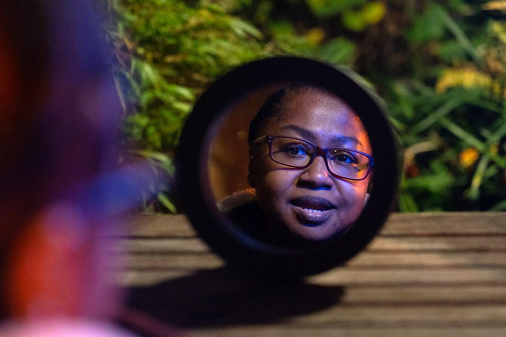 Jamilla McDaniel is seen through a reflection in a circular mirror that rests on a picnic table, her face is colored by red and blue light