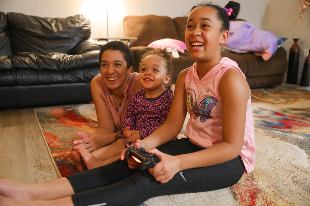 A woman and her two daughters smile as they sit on the floor and play a video game