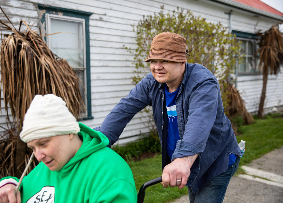 A man in a blue shirt and a bucket hat pushes a woman in a wheelchair, she is wearing a green shirt and white hat