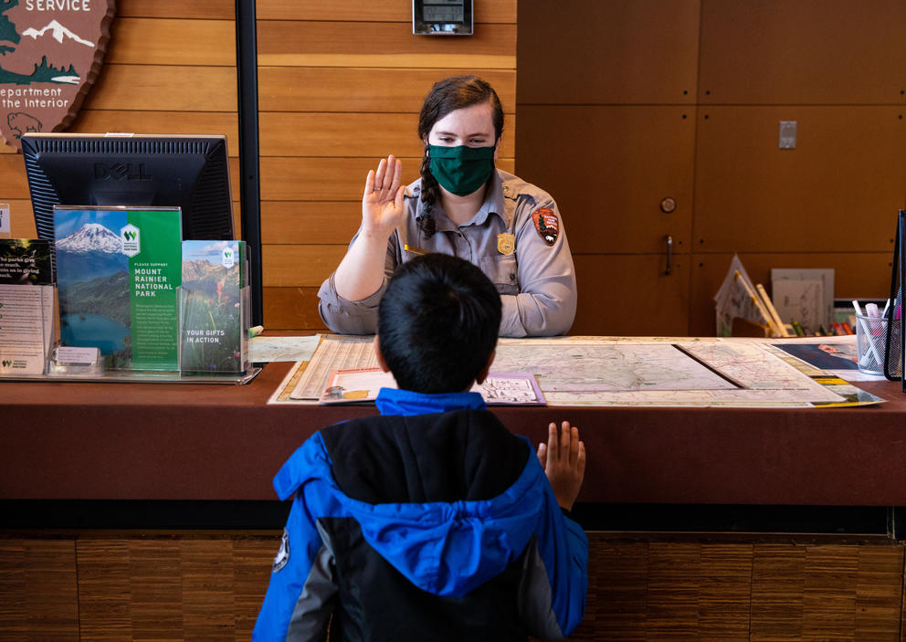 A boy with his back to the camera faces a park ranger sitting behind a desk. They both have their right hands raised.