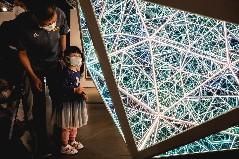 A small girl and her father look into a geometric sculpture filled with lines of LED lights