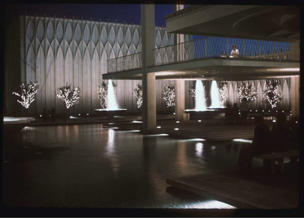 Night view of building made from white concrete. Lit-up fountains are spraying water in the air, and various platforms offer views of the mirror-like water below.