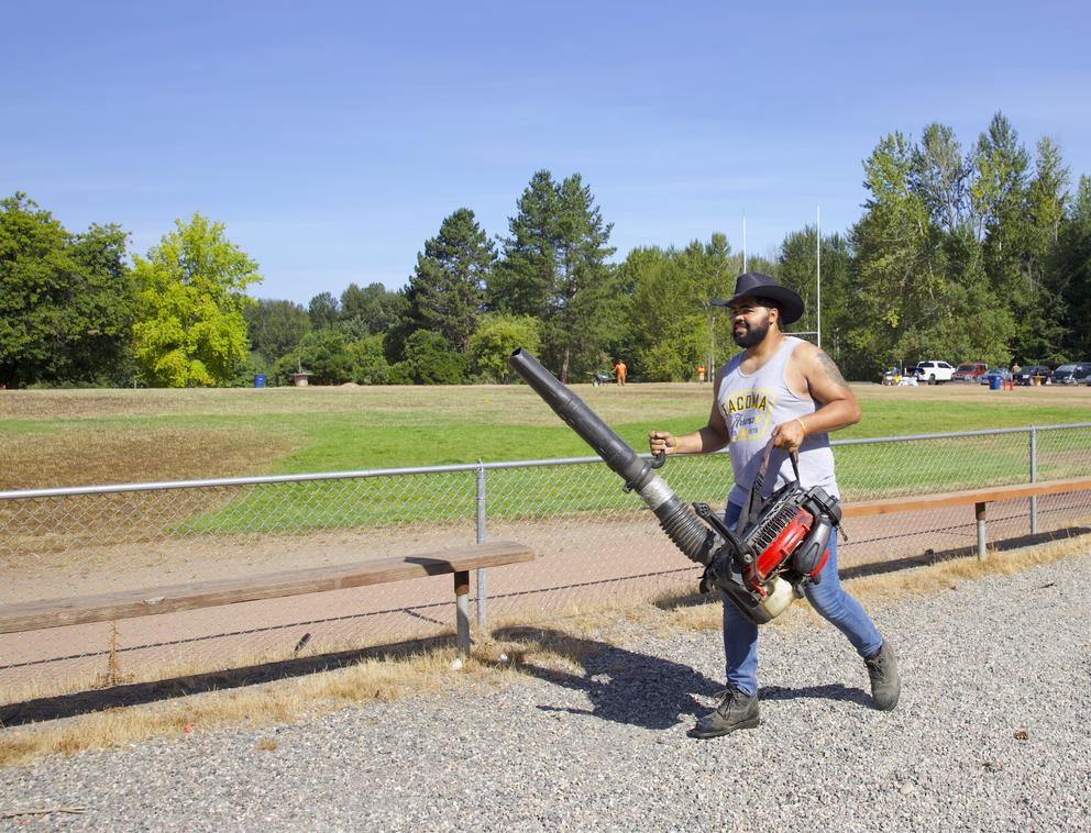 A person in a cowboy hat carries a leaf blower across a dirt path in the foreground of a ball field. There are trees in the background. 