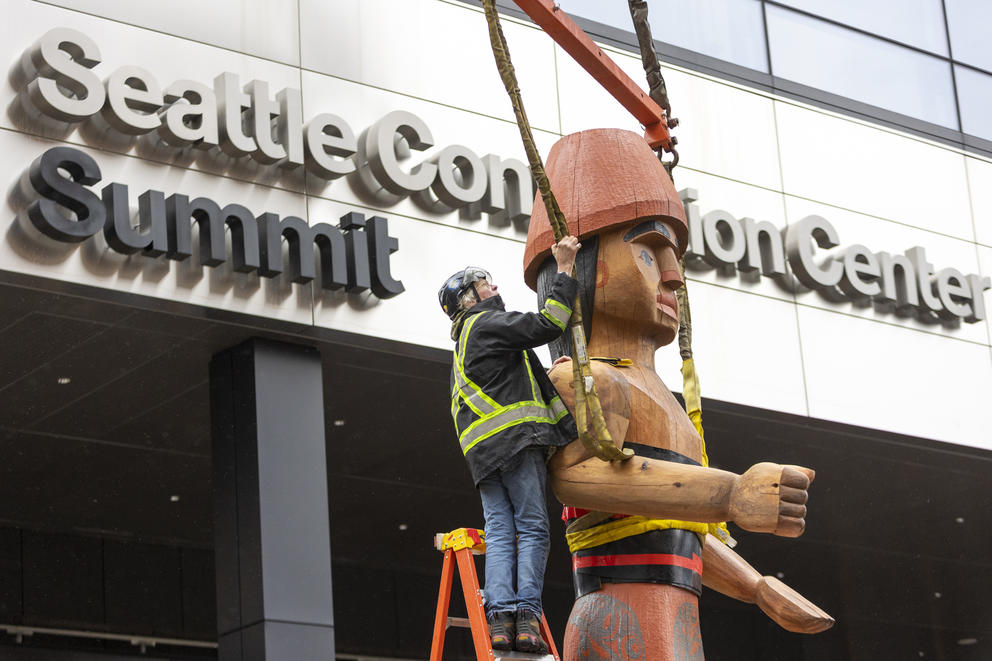 A worker stands on a ladder installing a large wooden sculpture in front of the convention center