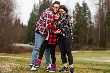 Sarah and Emily Colfer hug with their daughter between them. They are outdoor in a wooded area and wearing plaid.