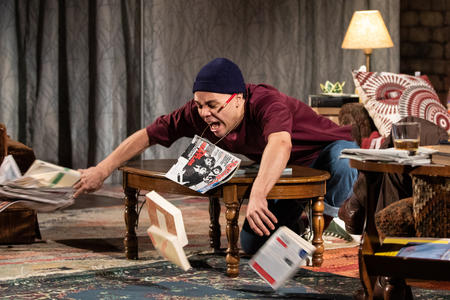 Person on a stage with lamp in the background throws magazines and books off a table, leaning over and with arms outstretched. They are wearing a bordeaux sweater.