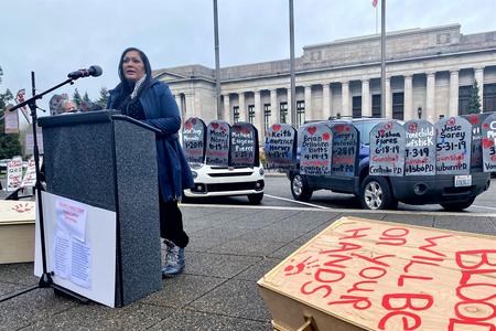 A woman stands outdoor at a podium at left, with a government building behind her. Also behind her are a string of paper signs in the shape of gravestones with names and dates on them. At right is a box shaped like a coffin that says, "Blood will be on your hands." 