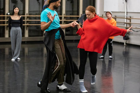 Person in blue t-shirt and black pants holds the hands of a person in black pants and red sweater as they are dancing and practicing steps in a rehearsal dance room