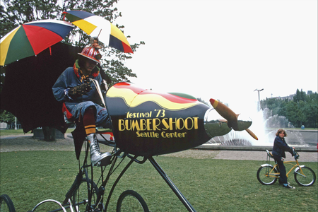 a vintage photo of a clownlike person riding a bike-plane combo with umbrellas attached