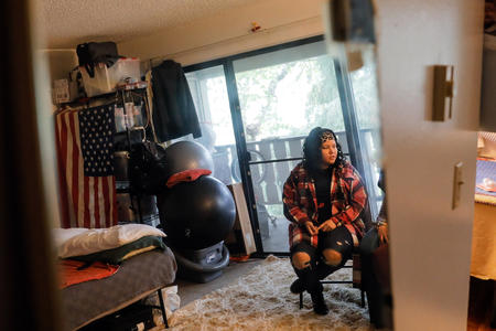 Janell Braxton sits in a folding chair wearing a flannel shirt while reflected in a mirror at her mother's apartment.