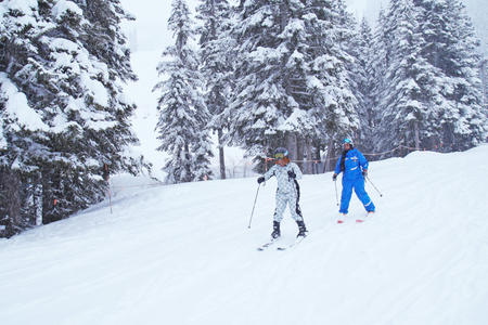 black woman in white snowsuit and black woman in blue ski down snowy slope surrounded by snow filled pine trees