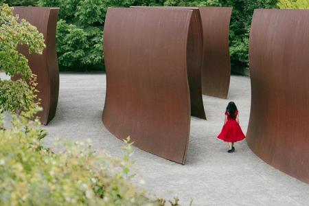 photo of a large sculptural outdoor installation that looks like rust colored waves, a woman in a red dress walks thru