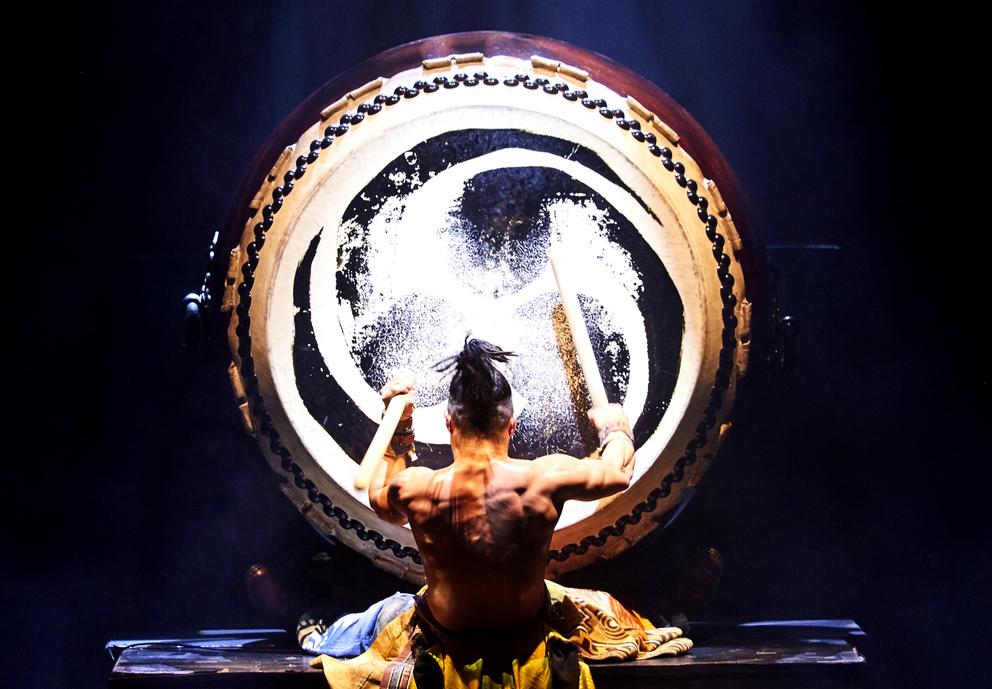 A man faces a large drum and plays it on stage