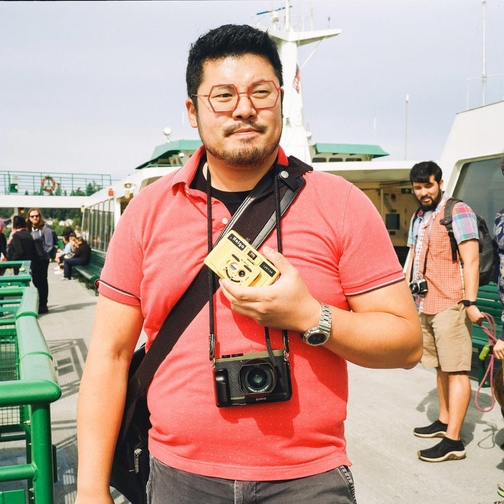 A man in a red shirt stands on a ferry deck with an analog camera around his neck and another in his hand