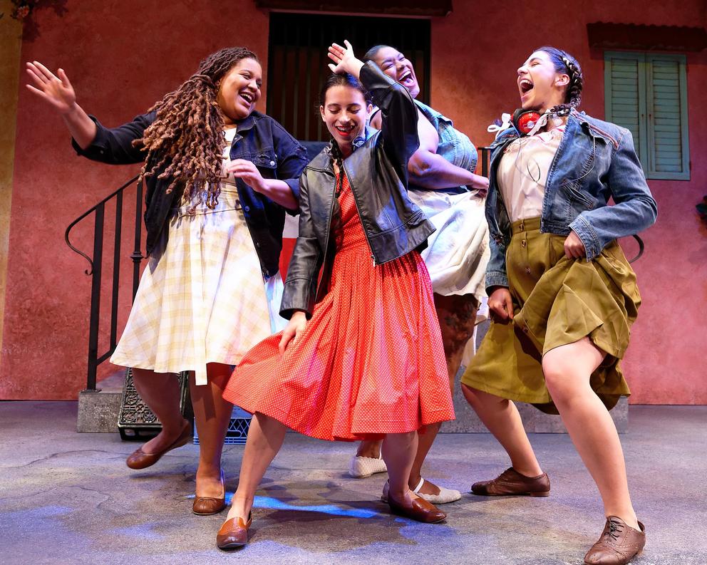 Four women in skirts are dancing in a group, having fun. They are on a stage with a (muted) red backdrop that looks like the façade of a house with iron staircase.