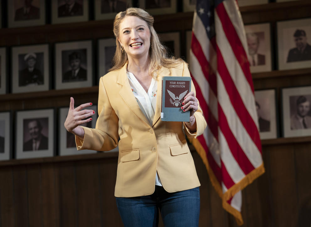 person in yellow blazer holds a book that says "Your rugged constitution", a U.S. flag hangs behind her