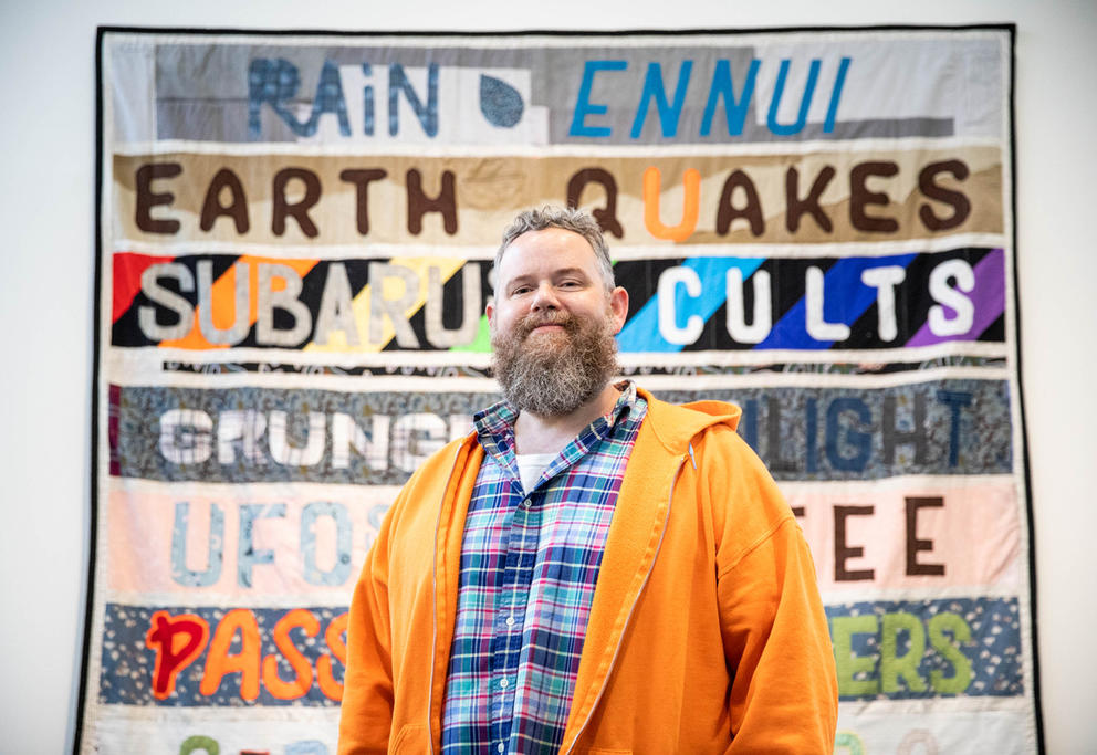 Portrait of a person with gray beard in front of a quilt with text reading RAIN ENNUI EARTH QUAKES and more