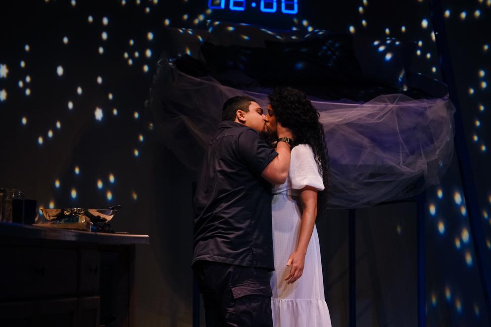 On a dark stage with lights, like stars, lit up, a man dressed in dark clothes kisses a woman in a white dress, her arms are besides her, he frames her face with his hands.