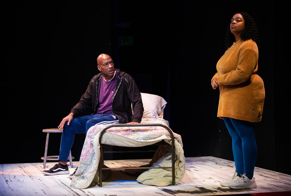 Two people on stage, one person - a man - sits on bed (left) while, to the right, a woman stands up straight. 