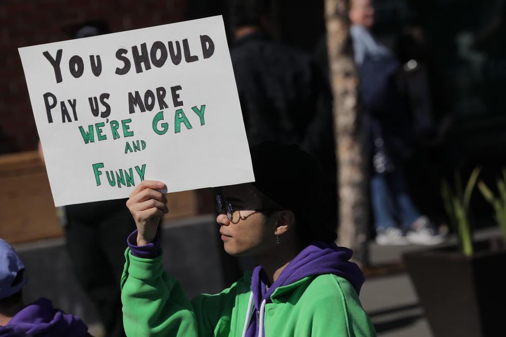 A man holds a sign reading "you should pay us more, we're gay and funny"