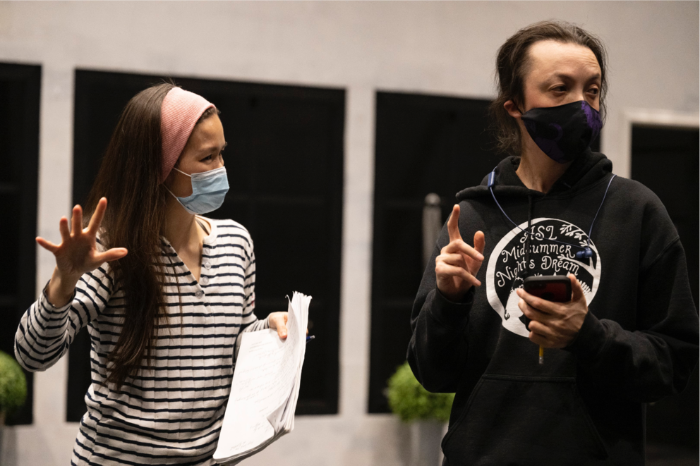 A woman wearing a mask gestures and looks at a man, also wearing a mask, to her right