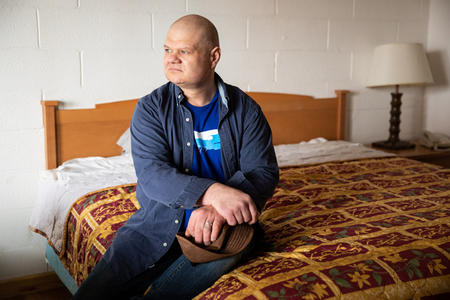 Jason Young sits on a hotel room bed, looking to his right, off camera, hands crossed in his lap.