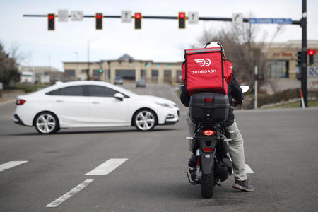 a doordash delivery person sits on a moped with a red food delivery bag on their back at a stop light on the street