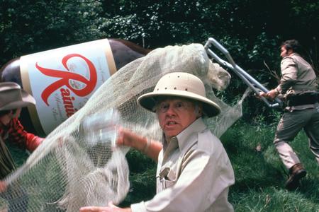 film still of an older man in a classic safari suit attempting to capture a giant Rainier Beer in a net